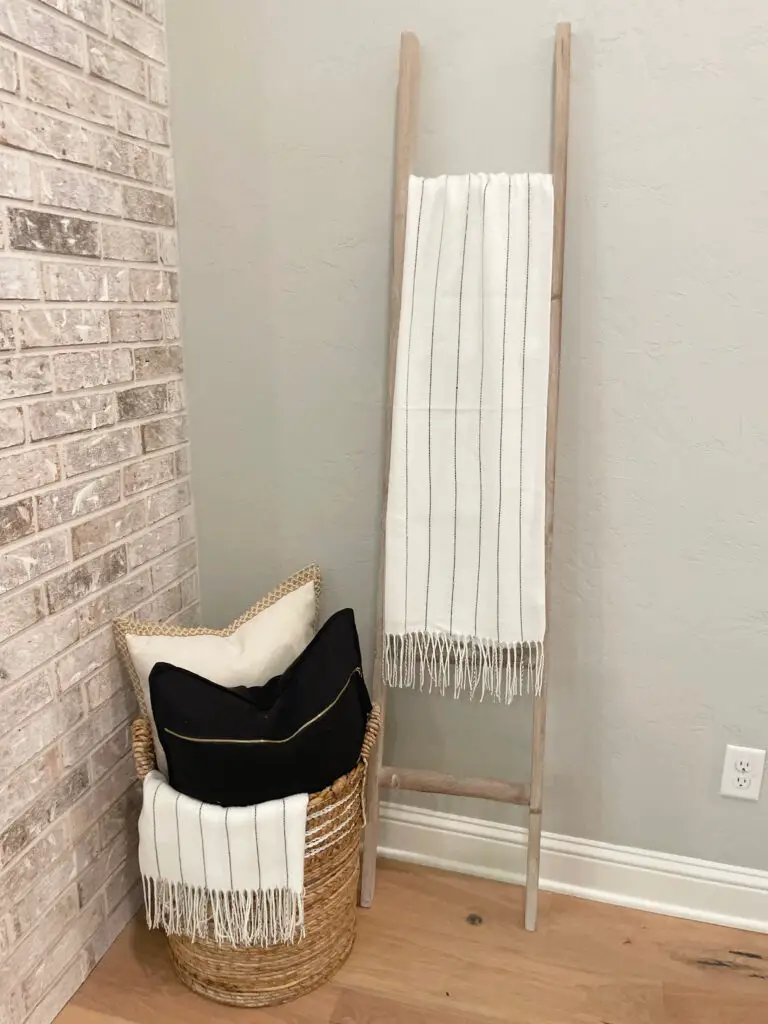 Pale wood ladder with white linen blanket styled next to a wicker basket filled with black and white linens & pillows
