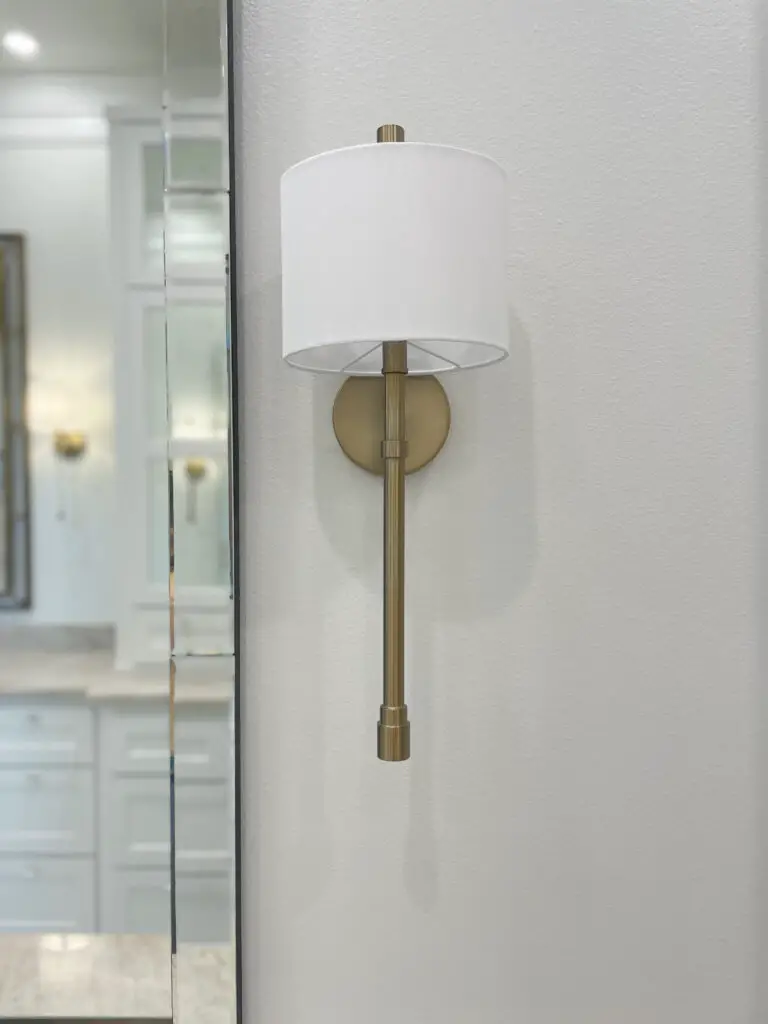 Gold stick wall sconce with flat round white shade next to mirror