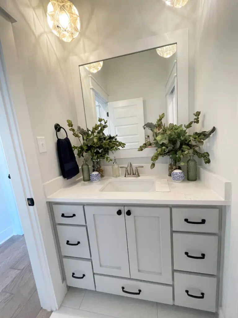 light grey cabinetry base of vanity with white counters, black hardware and green and blue accent decor in half bathroom
