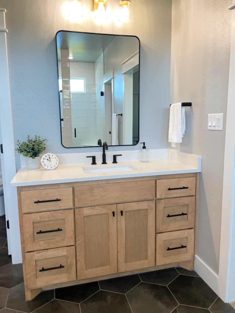 light wood vanity cabinets in master bathroom with white counter and green, white, and grey accents with black hardware