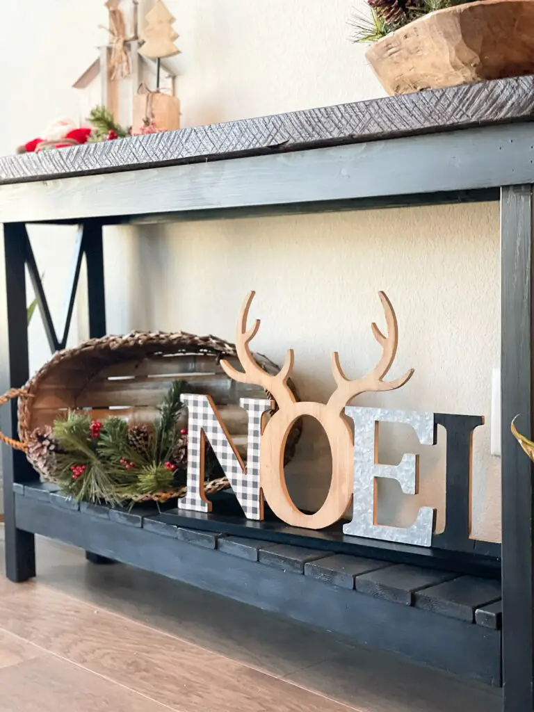 old basket on bottom of side table filled with a few Christmas greens behind a Noel sign